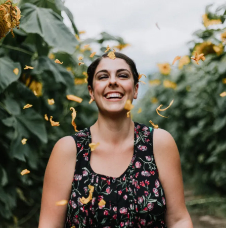A joyful woman with her head tilted back laughs as yellow petals fall around her in a field of tall sunflowers.